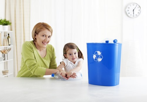A woman with short blonde hair and a young girl with pigtails are seated at a white kitchen or utility room countertop, engaging with a blue recycled plastic bin with a recycling symbol printed in white on its front. The woman is wearing a light green sweater and smiling at the camera, while the girl, dressed in a white top, looks slightly to the side with a neutral expression. The blue bin is positioned centrally on the counter and appears to be empty, with a lid resting on top. The background features a white wall, a wall clock showing approximately 1:02, and a wooden shelf holding small plants and decorative items. The environment suggests a clean and organized domestic setting, and the image visually supports waste management and recycling themes relevant to rubbish removal services in the Addlestone area near Surrey, with a focus on environmentally conscious disposal or recycling practices.