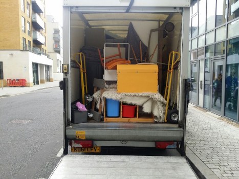 The rear of a small commercial van parked on a paved street in Addlestone, showing its open cargo area filled with various household items and rubbish. Inside the vehicle, there are two orange plastic chairs leaning against the back wall, a yellow cabinet or storage box placed near the centre, and a pile of miscellaneous waste materials including a black plastic bag, a small blue plastic storage bin, and a folded grey blanket or rug draped over some objects. The cargo area appears partially cluttered, with a few tools or equipment visible on the floor. The van’s exterior is grey, and the vehicle features a British-style yellow number plate. The background shows a modern urban setting with residential apartment buildings on the left and a glass-fronted commercial building on the right, typical of Addlestone town centre. This image relates to rubbish removal services offered by Commercial Waste Addlestone, focusing on domestic and small-scale waste collection and disposal in the local area.