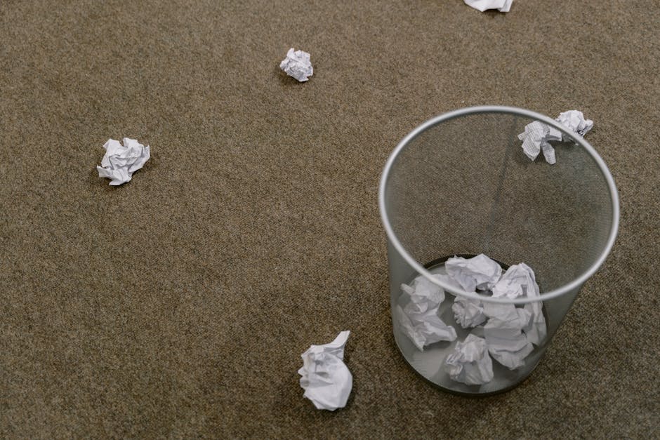 An empty cylindrical waste paper bin made of transparent plastic with a smooth, matte finish, positioned on a beige-brown textured carpeted floor. The bin is partially filled with crumpled white paper