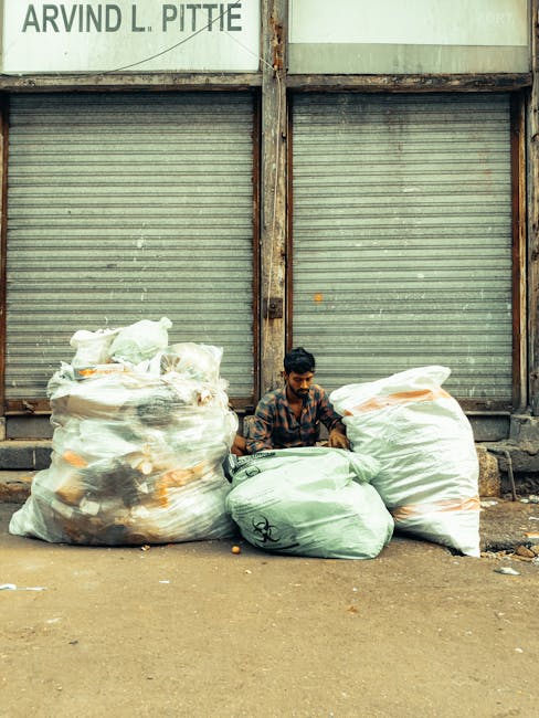 A large outdoor collection of waste materials consisting of multiple stacks of cardboard boxes, primarily filled with fresh fruits, located behind a row of green and red plastic waste bins. The cardbo
